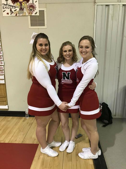 Three Navajo cheerleaders, standing in a pose, (about to launch the middle one onto the shoulders of the other two), in the corner of the Navajo Fieldhouse.